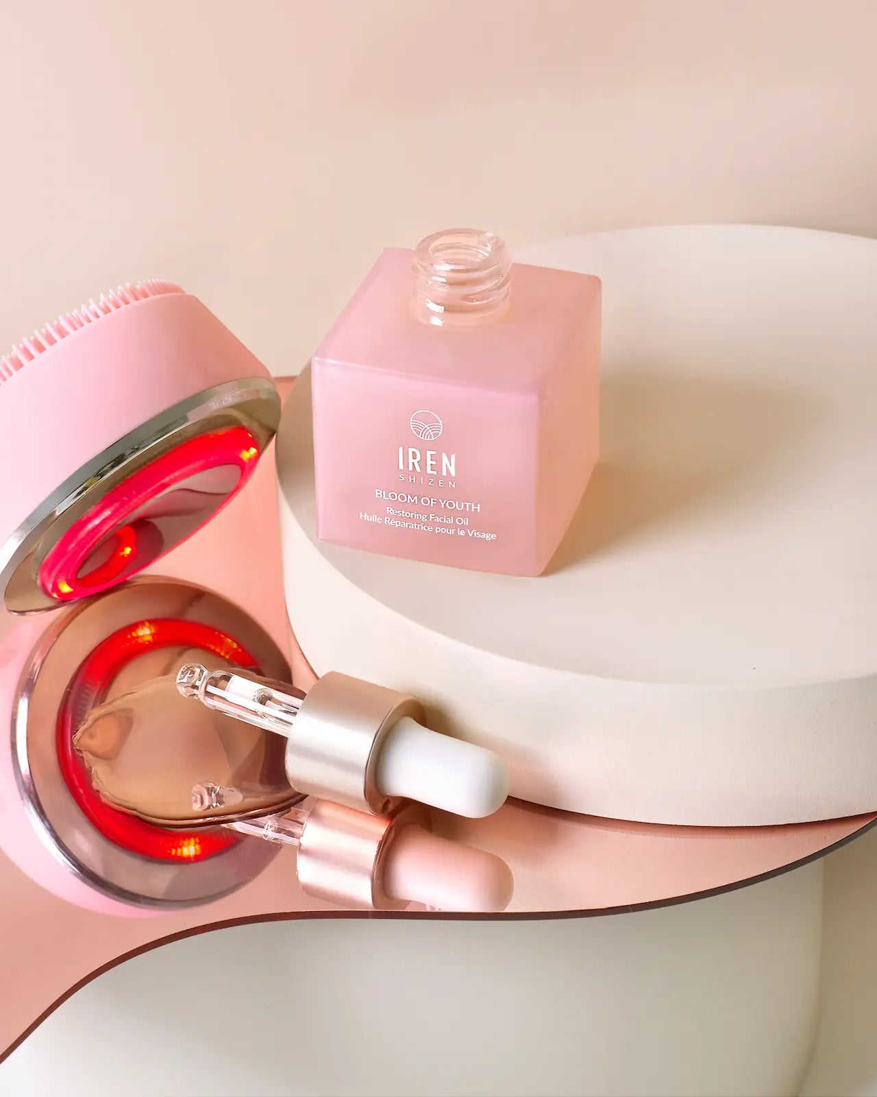 The GLOW ON THE GO Travel Light Kit by IREN Shizen features a pink facial cleansing brush, the Bloom of Youth square pink bottle, and a dropper—neatly arranged on mirrored platforms for your travel facial care needs.