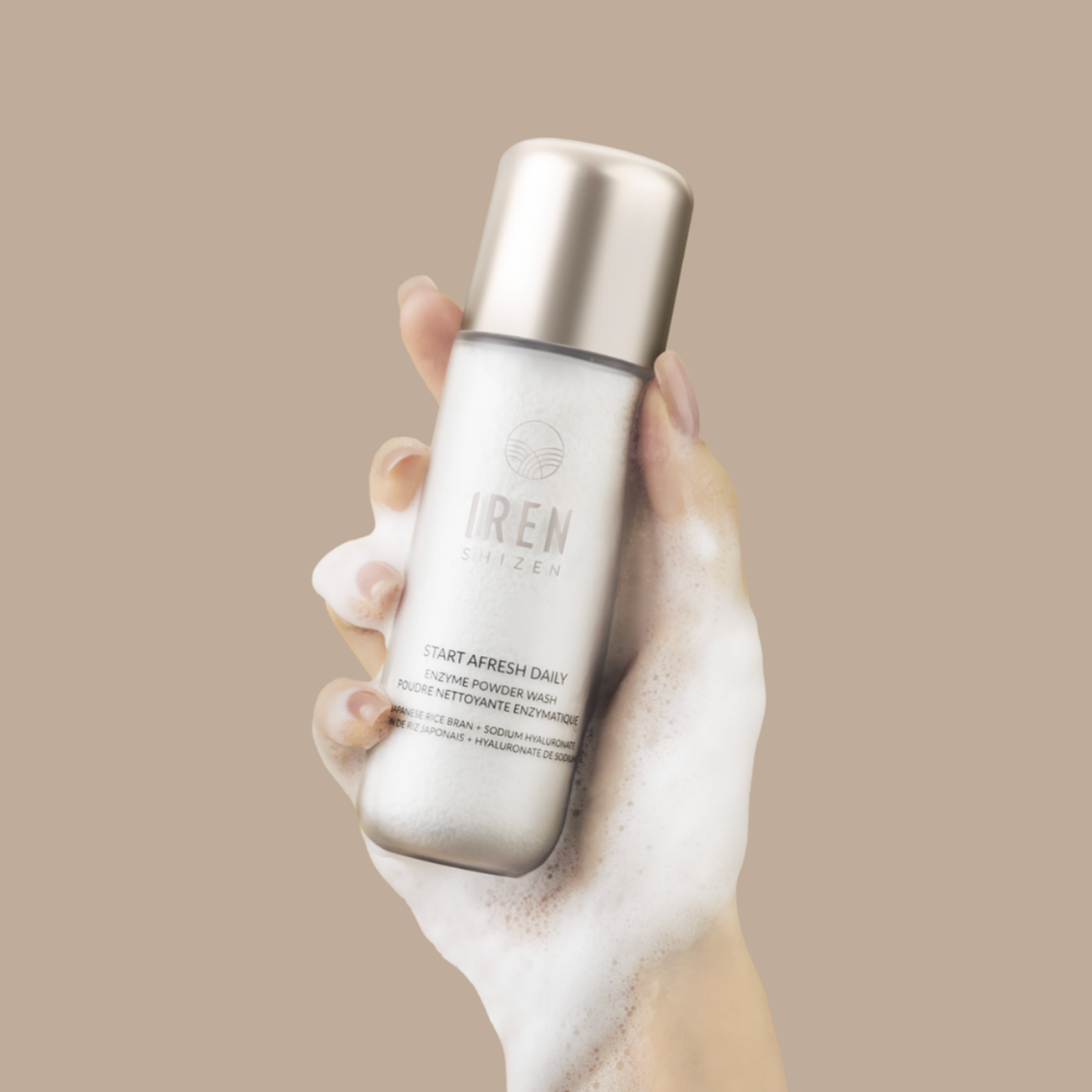 A hand covered in foam holds a bottle of Fast Bundle Cloud Cleansing Kit, a gentle enzyme cleanser, against a beige background.