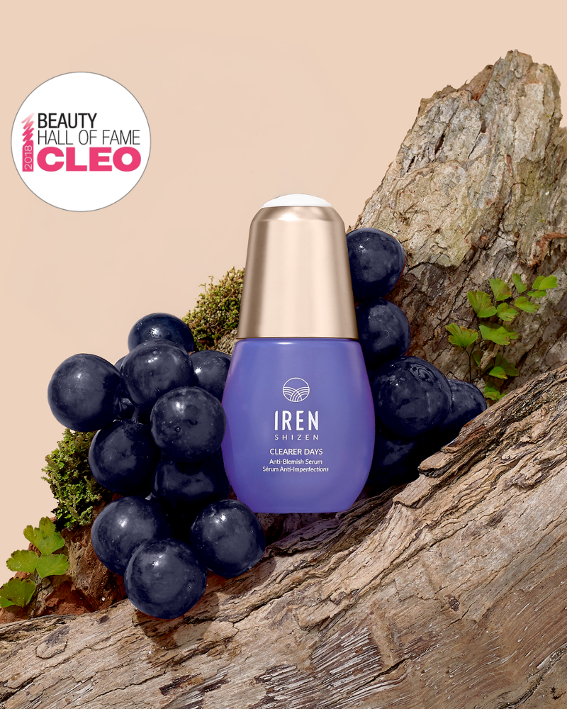 A bottle of IREN Shizen CLEARER DAYS Anti-Blemish Serum sits among grapes and tree bark, featuring a "Beauty Hall of Fame CLEO" badge in the top left corner.