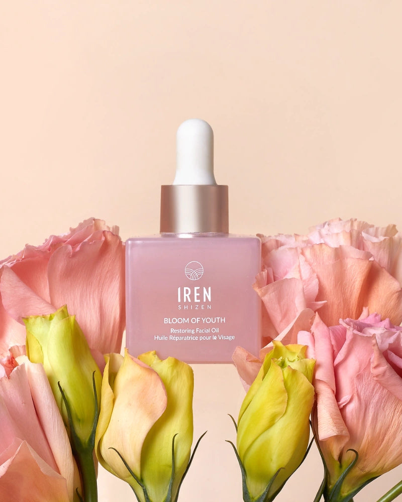 A pink dropper bottle labeled "BLOOM OF YOUTH Restoring Facial Oil" by IREN Shizen sits among pink and yellow flowers against a light beige background, highlighting its lightweight formula that supports skin barrier repair. #all-groups