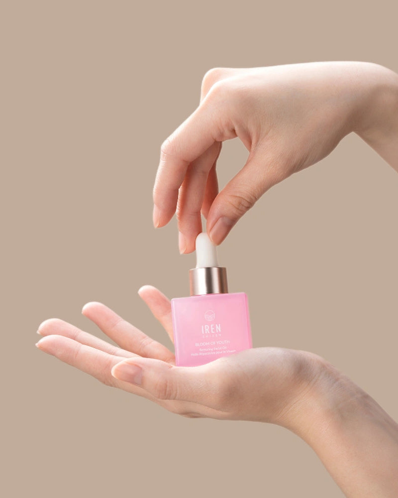 Someone holds a pink BLOOM OF YOUTH Restoring Facial Oil bottle from IREN Shizen in one hand and uses the dropper with the other against a beige background, showcasing skin barrier repair in their skincare routine.
#all-groups