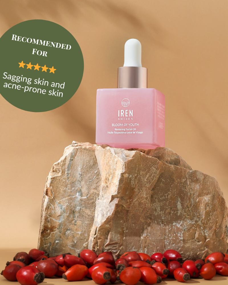 A pink bottle of BLOOM OF YOUTH Restoring Facial Oil by IREN Shizen with a dropper sits on a rock among rosehips; its green label highlights prebiotics and postbiotics for supporting the skin barrier, targeting sagging and acne-prone skin.