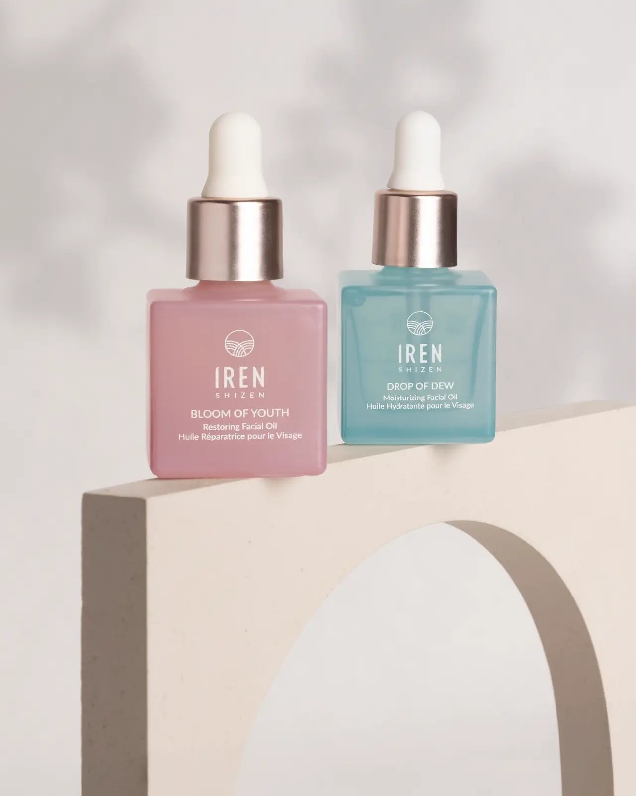 Two square IREN Shizen bottles, one pink and one blue, are displayed on a curved beige stand next to the GLOW ON THE GO Travel Light Kit, set against a neutral background.