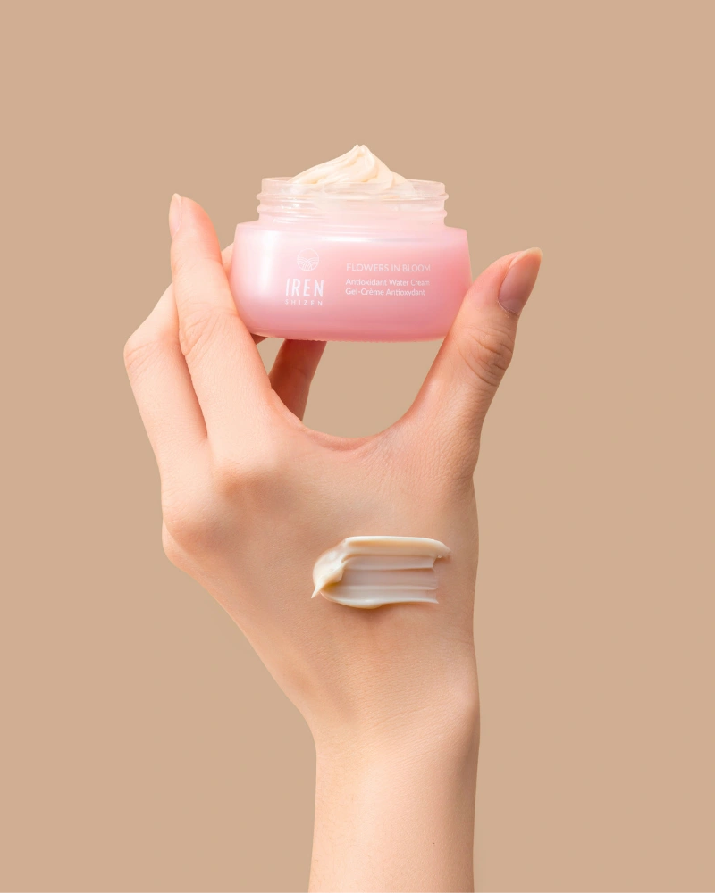 A hand holds an open jar of IREN Shizen FLOWERS IN BLOOM Antioxidant Water Cream against a beige background, with a swipe of the hydrating formula applied to the back of the hand. #all-groups