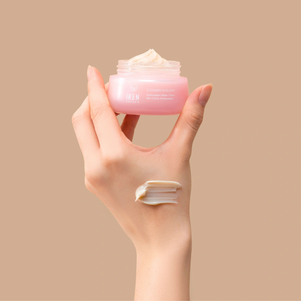 A hand holds an open jar of IREN Shizen FLOWERS IN BLOOM Antioxidant Water Cream against a beige background, with a swipe of the hydrating formula applied to the back of the hand. #all-groups