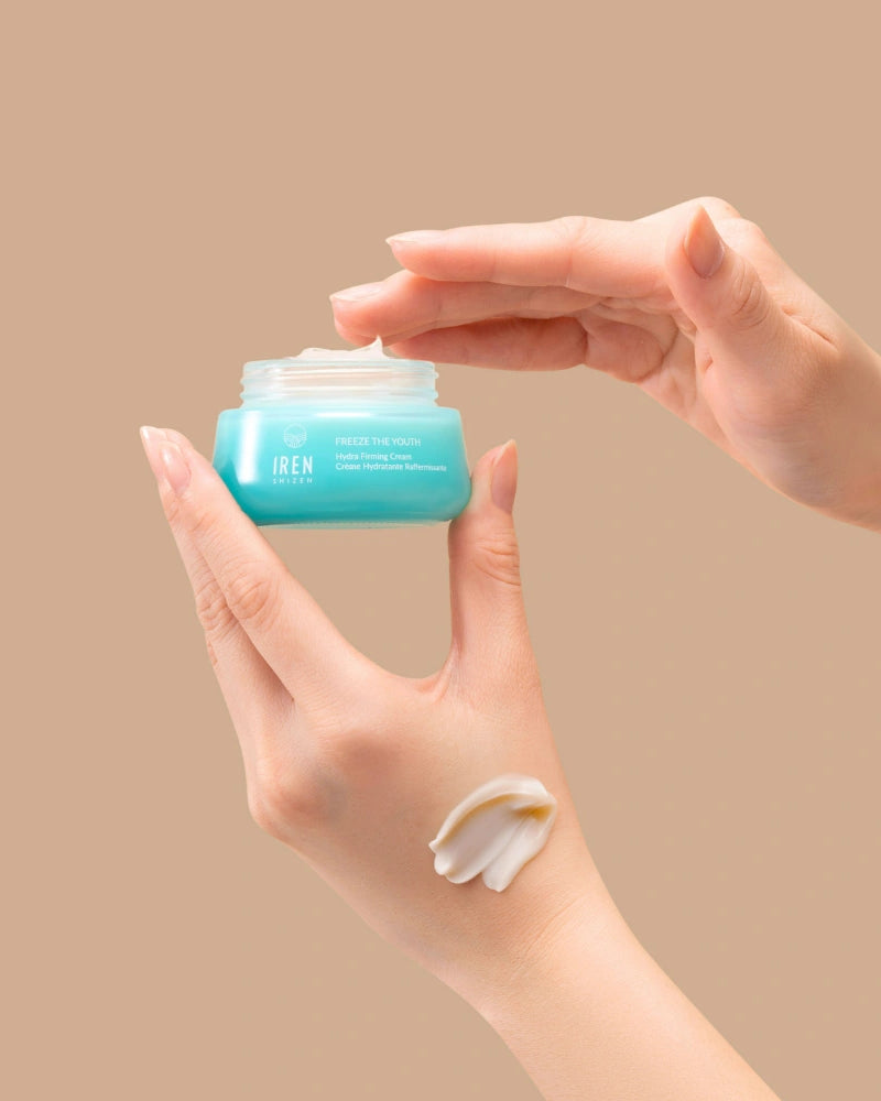 A hand holds an open jar of IREN Shizen FREEZE THE YOUTH Hydra Firming Cream while the other dips in; a smear of this hydrating, age-defying moisturizer is visible on the wrist against a beige background. #all-groups
