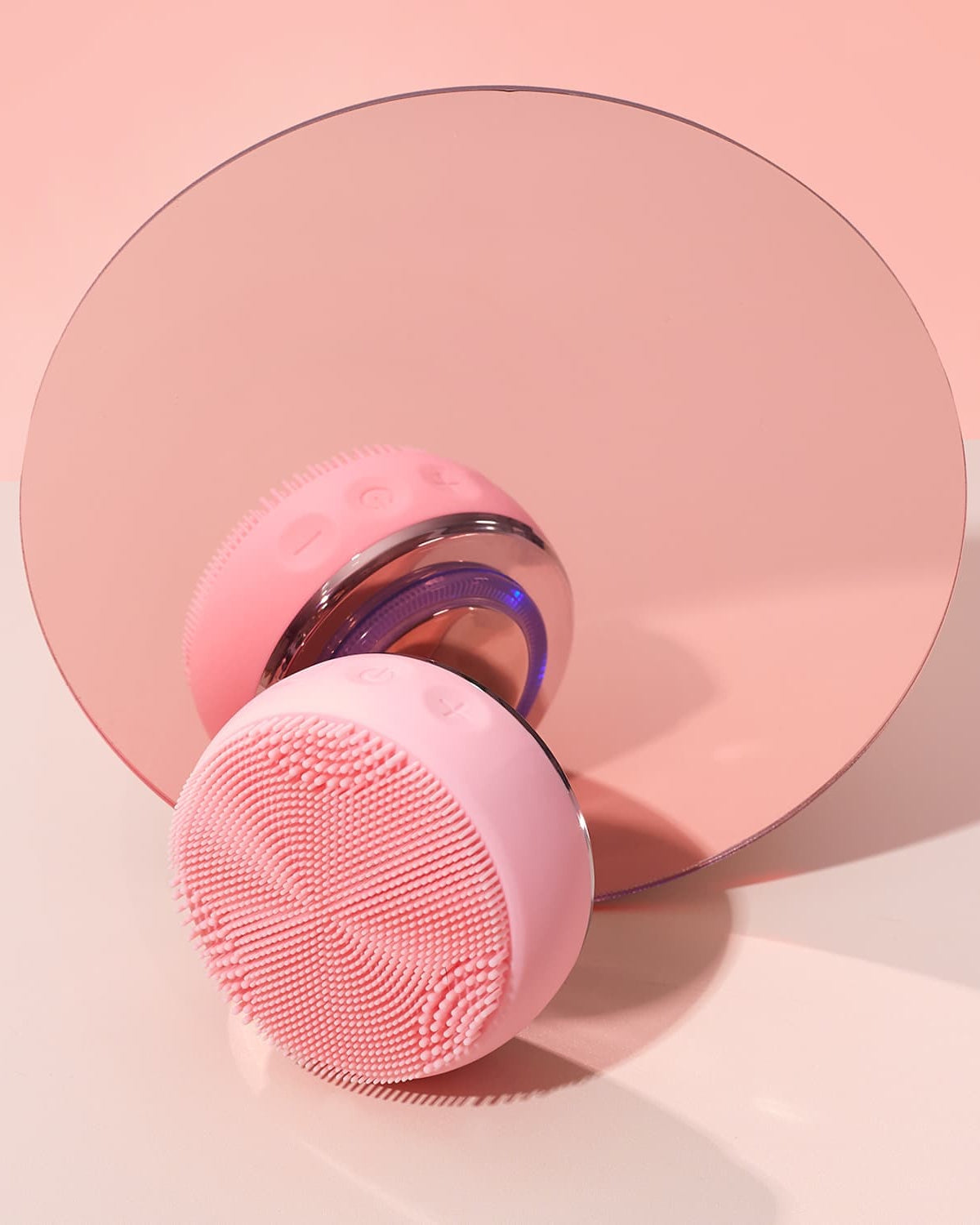 The IREN Shizen GLOW ON THE GO Travel Light Kit's pink silicone facial cleansing device rests before a round mirror on a light pink background—ideal for your travel skincare routine.