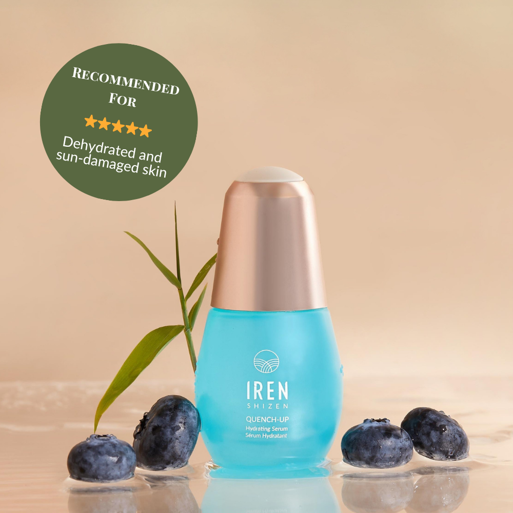 A blue bottle of IREN Shizen QUENCH-UP Hydrating Serum with hyaluronic acid sits on a wet surface among blueberries and a green leaf, labeled for dehydrated and sun-damaged skin to help achieve plump skin.