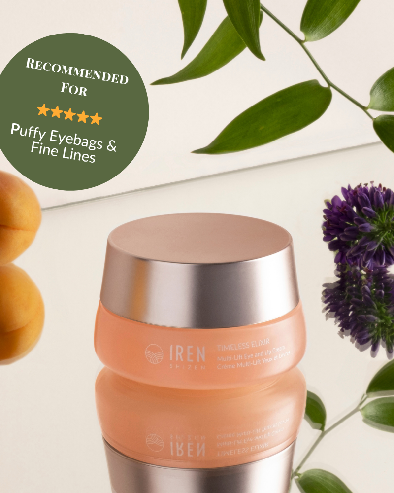 A peach IREN Shizen TIMELESS ELIXIR Multi-Lift Eye and Lip Cream jar sits on a reflective surface with leaves, purple flowers, and apricots. A green badge recommends it for treating puffy eyebags, fine lines, and dark circles.