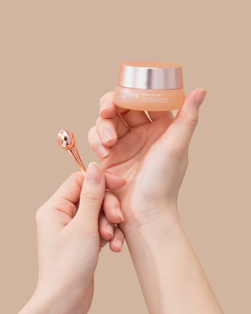 Hands holding a small jar of IREN Shizen TIMELESS ELIXIR Multi-Lift Eye and Lip Cream and a metal applicator against a beige background, perfect for eye area rejuvenation.