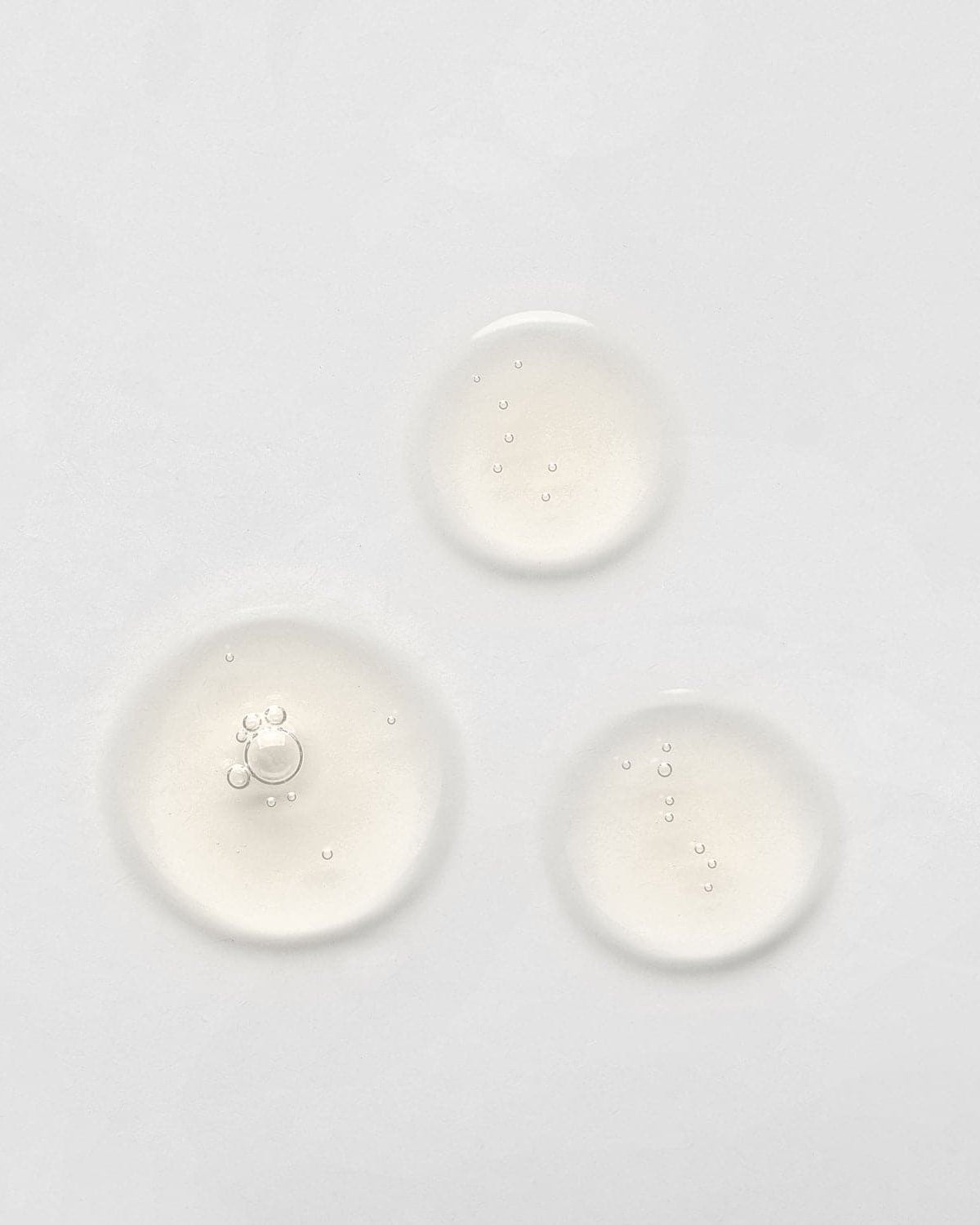 Three small, clear liquid droplets on a plain white surface, possibly from the LIFT & FIRM Anti-Aging Duo Serum Set by IREN Shizen, which contains Argireline Peptide. #all-groups