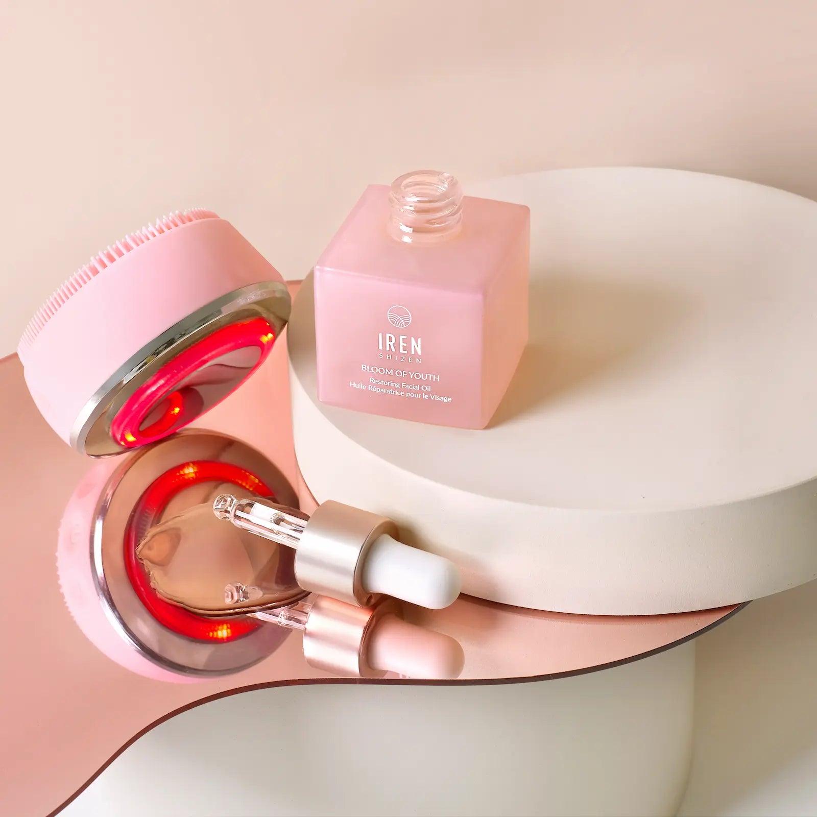 The GLOW ON THE GO Travel Light Kit by IREN Shizen features a pink facial cleansing brush, the Bloom of Youth square pink bottle, and a dropper—neatly arranged on mirrored platforms for your travel facial care needs.