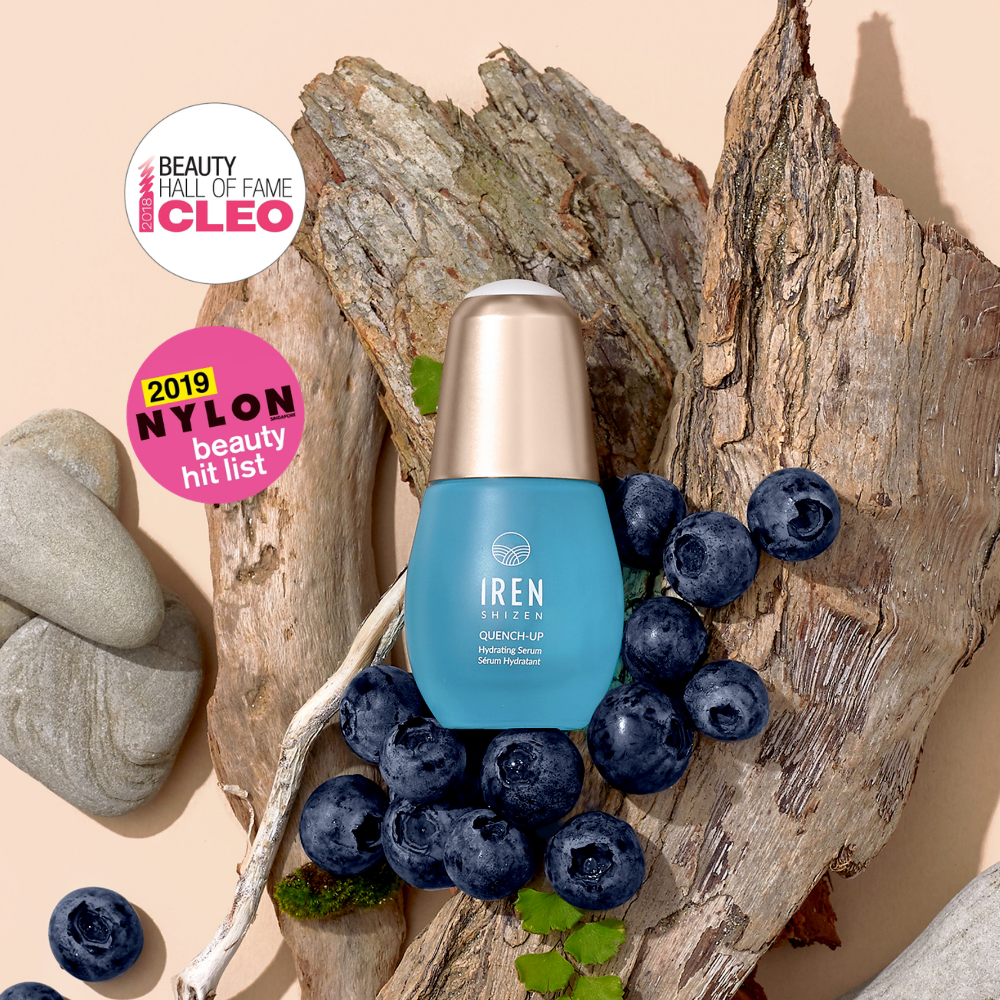 A blue QUENCH-UP Hydrating Serum bottle by IREN Shizen is surrounded by blueberries, driftwood, stones, and CLEO and NYLON award stickers—ideal for plump skin with Hyaluronic Acid.
