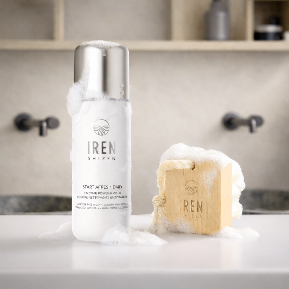 The Fast Bundle Cloud Cleansing Kit, featuring a gentle enzyme cleanser and a bubbly soap with a facial cleansing brush, sits on the bathroom counter; a blurred sink and shelves appear in the background.