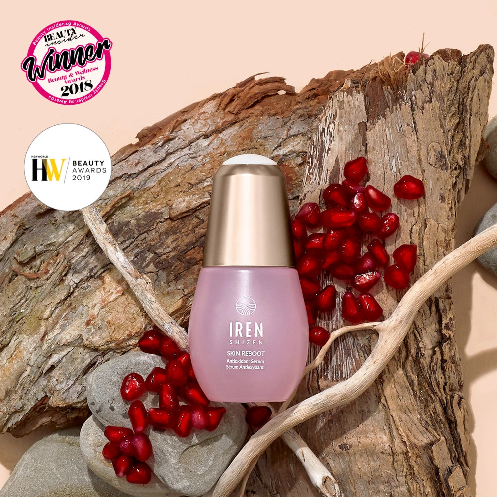 A bottle of IREN Shizen SKIN REBOOT Antioxidant Serum rests on wood with pomegranate seeds, rocks, and two beauty award badges, emphasizing its pomegranate extract for reducing dark circles.