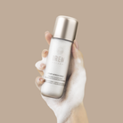 A hand covered in foam holds a bottle of Fast Bundle Cloud Cleansing Kit, a gentle enzyme cleanser, against a beige background.