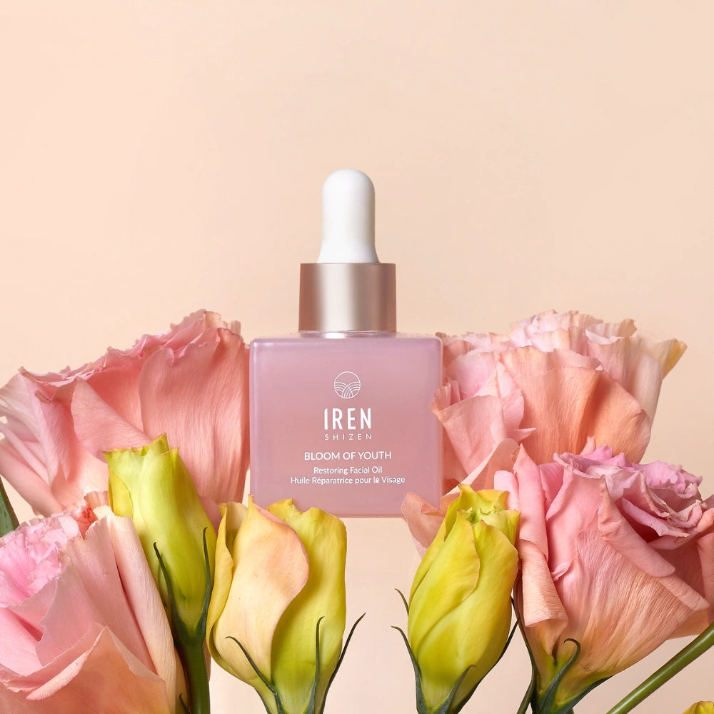 A pink dropper bottle labeled "BLOOM OF YOUTH Restoring Facial Oil" by IREN Shizen sits among pink and yellow flowers against a light beige background, highlighting its lightweight formula that supports skin barrier repair. #all-groups