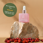 A pink bottle of BLOOM OF YOUTH Restoring Facial Oil by IREN Shizen with a dropper sits on a rock among rosehips; its green label highlights prebiotics and postbiotics for supporting the skin barrier, targeting sagging and acne-prone skin. #show-lang-en