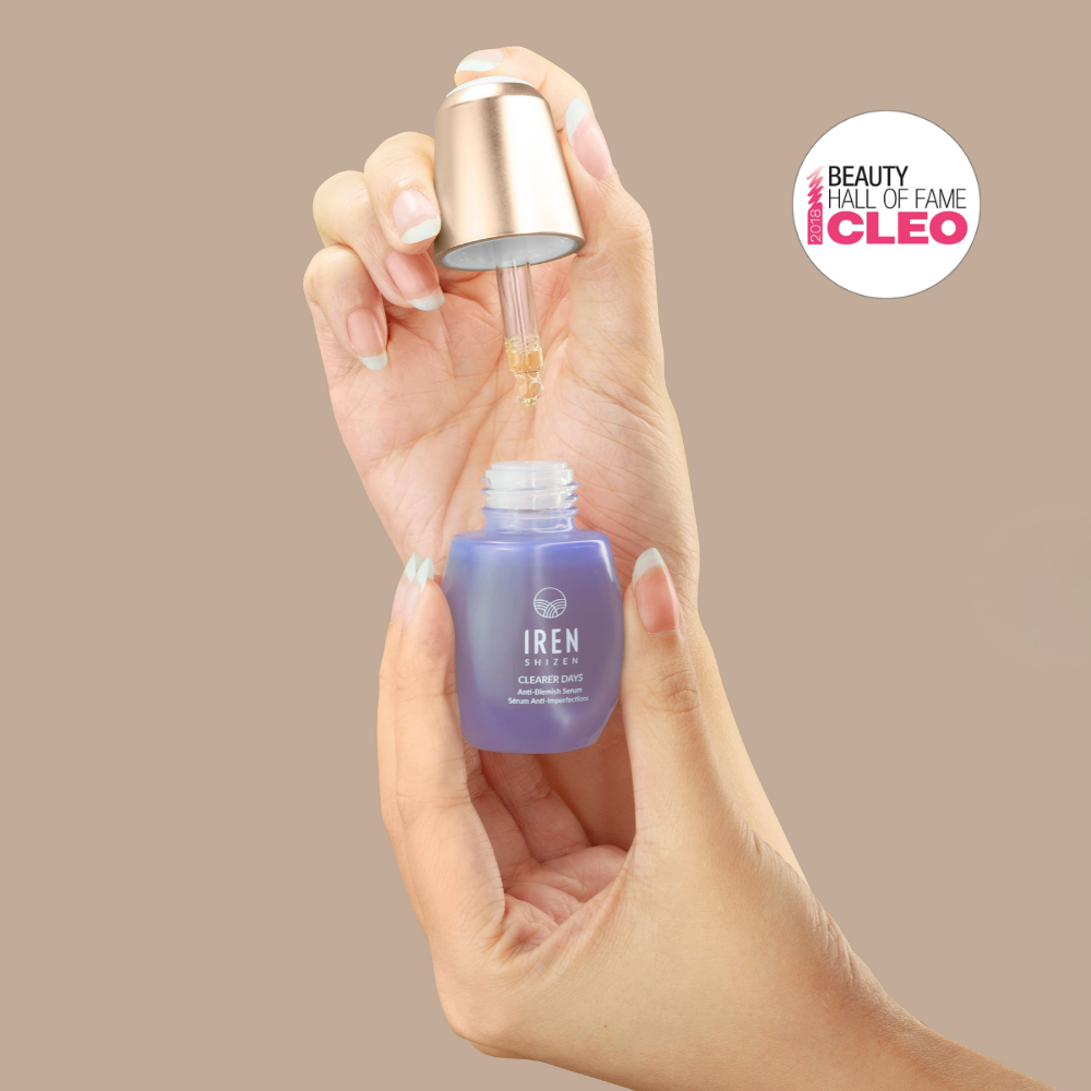 A hand holds an open bottle of IREN Shizen CLEARER DAYS Anti-Blemish Serum, dispensing liquid with a dropper. A "Beauty Hall of Fame CLEO" badge highlights the serum’s advanced acne treatment and sebum balance benefits.