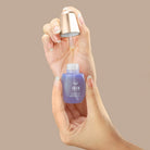 A hand holds a purple IREN Shizen CLEARER DAYS Anti-Blemish Serum bottle while another dispenses the serum with a dropper against a beige background, highlighting its acne treatment and sebum-balancing benefits. #all-groups