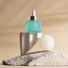 A blue GLOW ON THE GO Travel Light Kit by IREN Shizen balances on a broken stone slab with a white sphere and textured base, capturing travel elegance against a soothing beige background.