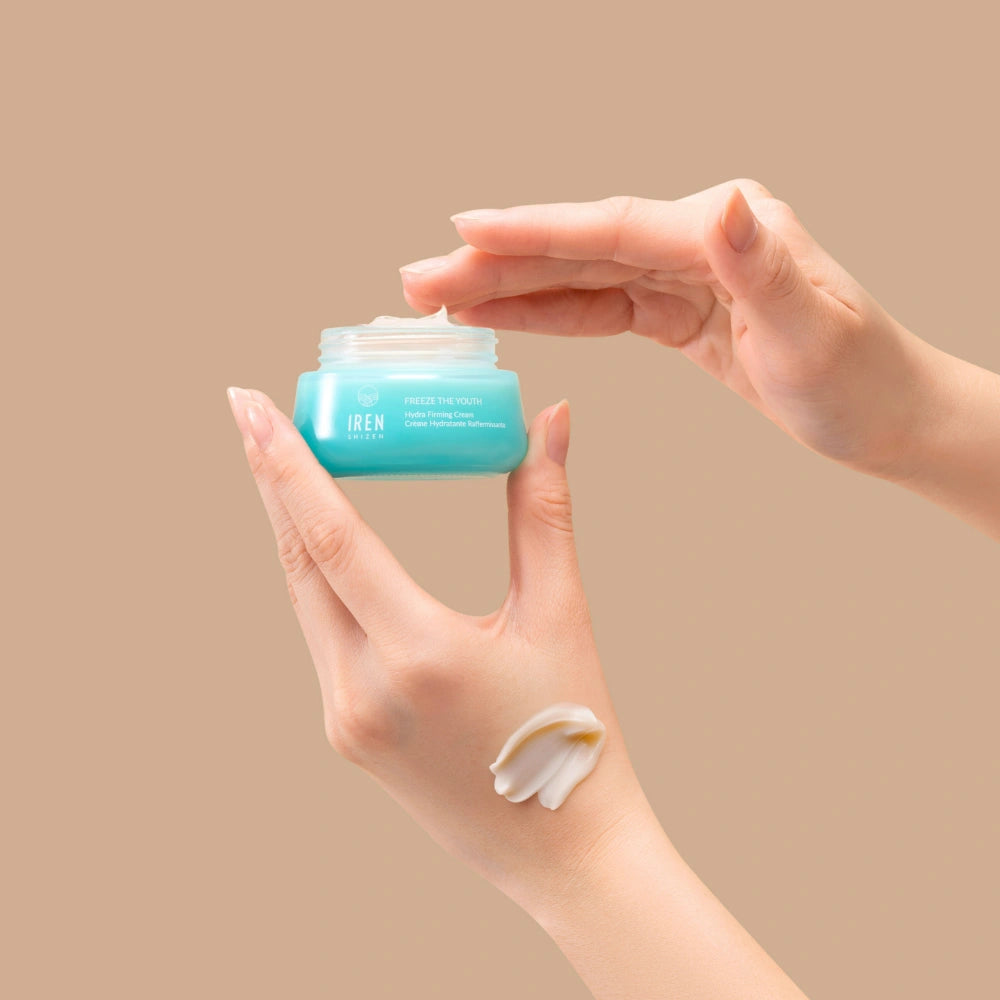A hand holds an open jar of IREN Shizen FREEZE THE YOUTH Hydra Firming Cream while the other dips in; a smear of this hydrating, age-defying moisturizer is visible on the wrist against a beige background. #all-groups
