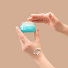 A hand holds an open jar of IREN Shizen FREEZE THE YOUTH Hydra Firming Cream while the other dips in; a smear of this hydrating, age-defying moisturizer is visible on the wrist against a beige background. #all-groups