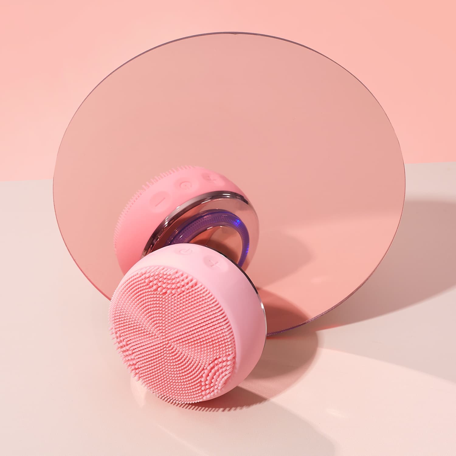 The IREN Shizen GLOW ON THE GO Travel Light Kit's pink silicone facial cleansing device rests before a round mirror on a light pink background—ideal for your travel skincare routine.