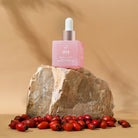 A pink dropper bottle labeled "IREN Shizen GLOW ON THE GO Travel Light Kit" sits on a rock, surrounded by scattered rose hips and a travel skincare kit against a beige background.
