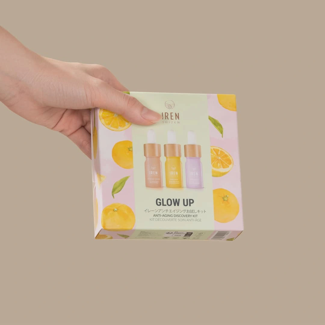 A hand holds a box of the IREN Shizen GLOW UP Anti-Aging Discovery Kit, featuring colorful citrus graphics and images of three serum bottles on the packaging.
