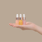 A hand holds three small dropper bottles from the IREN Shizen GLOW UP Anti-Aging Discovery Kit, featuring different colored serums against a plain beige background.