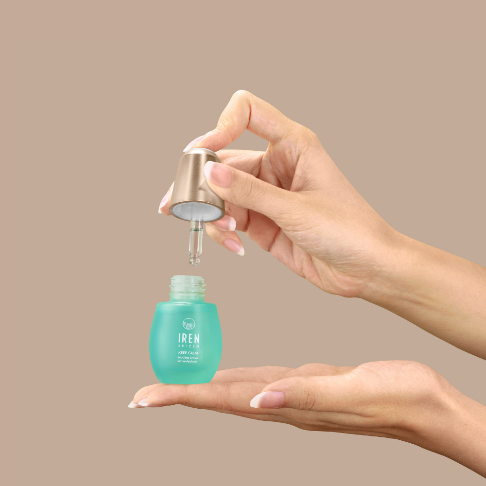 A hand dispenses KEEP CALM Soothing Serum by IREN Shizen from a blue bottle onto the palm, ideal for sensitive skin, with a plain beige background.