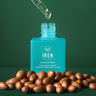 A dropper dispenses oil into a blue bottle labeled "IREN Shizen GLOW ON THE GO Travel Light Kit," surrounded by brown seeds on a green background—making it the perfect choice for any travel skincare routine.