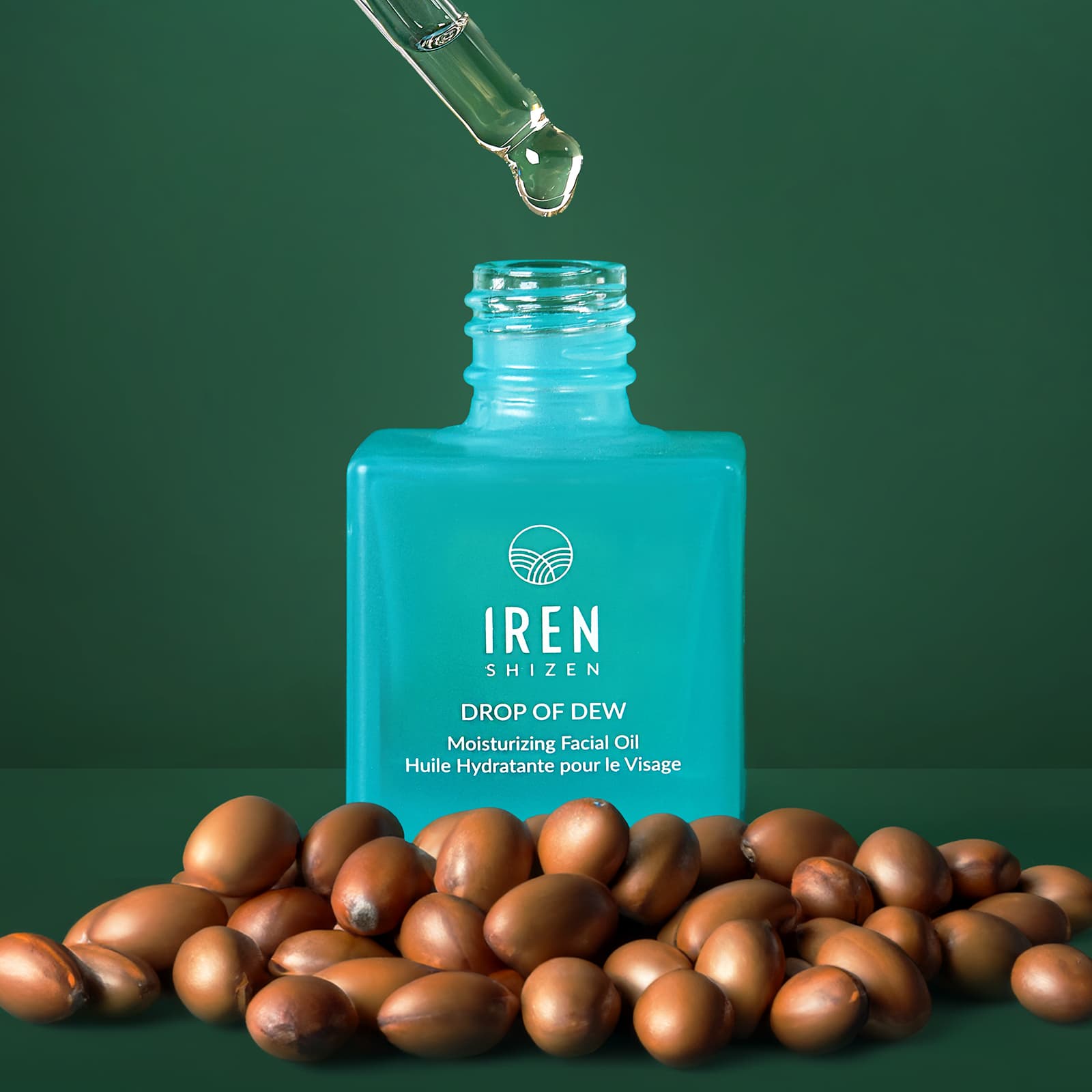 A dropper dispenses oil into a blue bottle labeled "IREN Shizen GLOW ON THE GO Travel Light Kit," surrounded by brown seeds on a green background—making it the perfect choice for any travel skincare routine.
