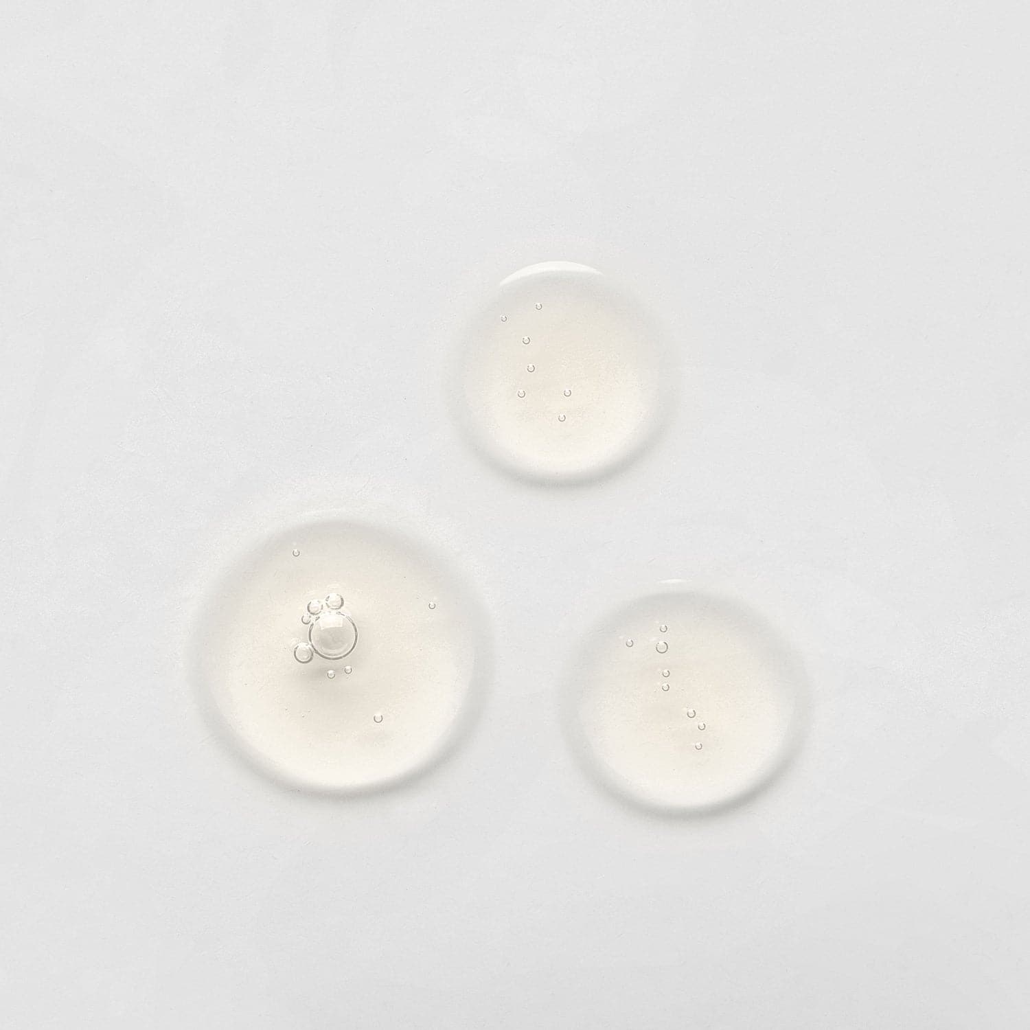 Three small, clear liquid droplets on a plain white surface, possibly from the LIFT & FIRM Anti-Aging Duo Serum Set by IREN Shizen, which contains Argireline Peptide. #all-groups